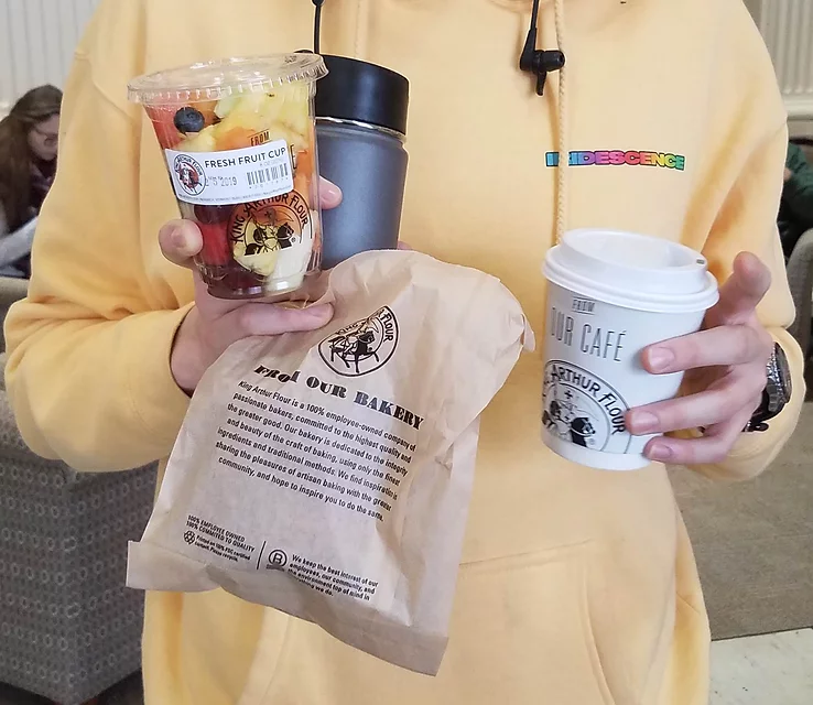 A close-up on a student holding a bagged pastry, a parfait, an iced-coffee, and a waterbottle.