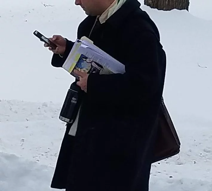 A professor with arms full of papers, books, yet carrying a bag, looks at his phone while walking.
